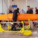 A team of technicians and engineers are assembling or inspecting a large orange unmanned aerial vehicle (UAV) or drone inside a hangar or tent facility. The aircraft is mounted on yellow wheeled support stands, and multiple personnel are working around it simultaneously. The setting appears to be a professional aerospace assembly or maintenance environment.
