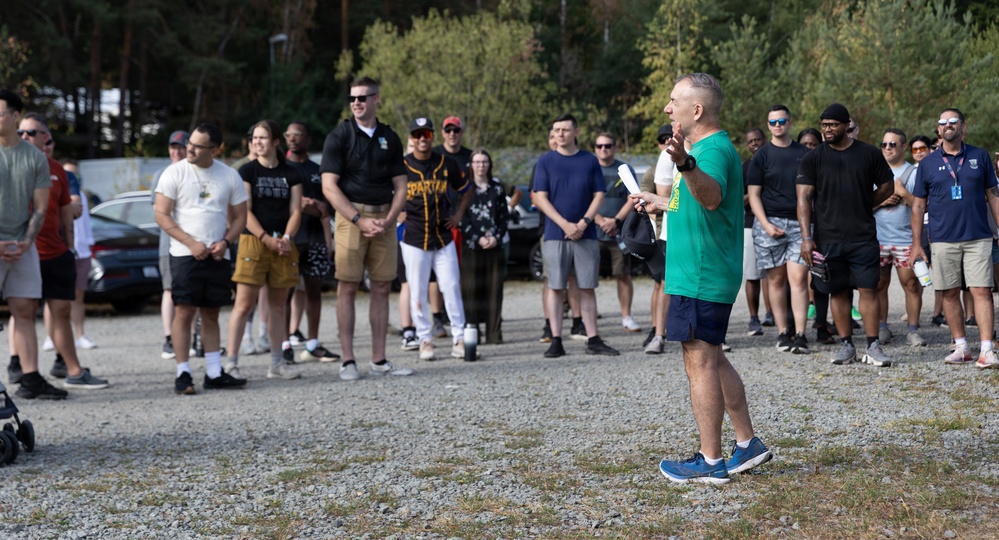 A man in a green shirt and blue shorts stands facing a large group of people in an outdoor gravel area, appearing to address or brief them while holding papers. The crowd of approximately 25-30 people is gathered in a semi-circle listening attentively. The setting appears to be a parking or staging area with trees in the background, suggesting an outdoor event or activity briefing.