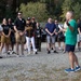 A man in a green shirt and blue shorts stands facing a large group of people in an outdoor gravel area, appearing to address or brief them while holding papers. The crowd of approximately 25-30 people is gathered in a semi-circle listening attentively. The setting appears to be a parking or staging area with trees in the background, suggesting an outdoor event or activity briefing.