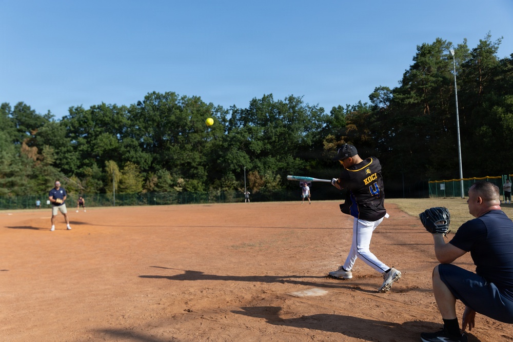 A softball game is in progress on a dirt infield field surrounded by trees under a clear blue sky. A batter wearing jersey number 4 with the name 'KOCI' has just swung at a yellow softball, which is visible in the air. A catcher crouches to the right while a fielder stands in the background near the pitcher's mound area.