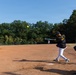 A softball game is in progress on a dirt infield field surrounded by trees under a clear blue sky. A batter wearing jersey number 4 with the name 'KOCI' has just swung at a yellow softball, which is visible in the air. A catcher crouches to the right while a fielder stands in the background near the pitcher's mound area.