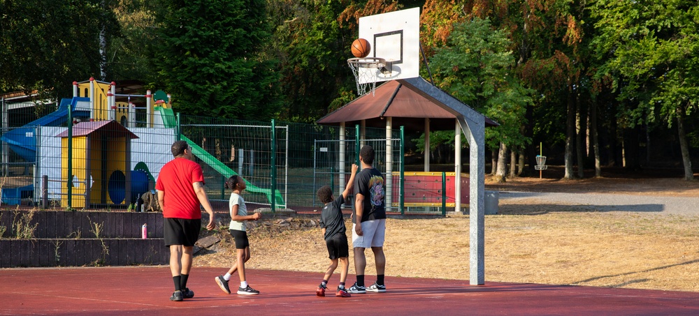 A group of four people, including two adults and two children, are playing basketball on an outdoor red court. The scene appears to be a family or group recreational activity, with a basketball visible near the hoop mid-shot. The court is situated next to a colorful playground area surrounded by green trees.