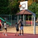 A group of four people, including two adults and two children, are playing basketball on an outdoor red court. The scene appears to be a family or group recreational activity, with a basketball visible near the hoop mid-shot. The court is situated next to a colorful playground area surrounded by green trees.