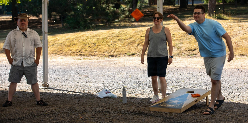 Three people are playing cornhole (bean bag toss) outdoors on a gravel surface, likely at a park or recreational area. A person in a light blue shirt has just thrown an orange bean bag, which is visible mid-air. The cornhole board is branded with a U.S. Army logo.