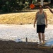 Three people are playing cornhole (bean bag toss) outdoors on a gravel surface, likely at a park or recreational area. A person in a light blue shirt has just thrown an orange bean bag, which is visible mid-air. The cornhole board is branded with a U.S. Army logo.