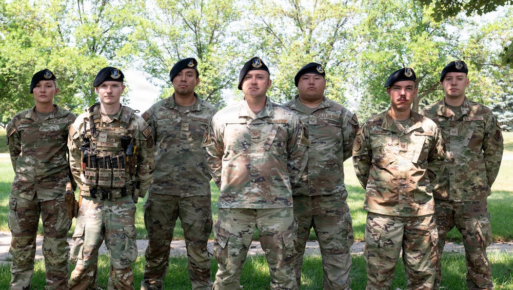 Seven U.S. Air Force personnel in OCP (Operational Camouflage Pattern) uniforms are posed together outdoors in a group photo. All individuals are wearing black berets with what appears to be the U.S. Space Force or Air Force insignia. The setting is a grassy, tree-lined area, likely on a military installation.