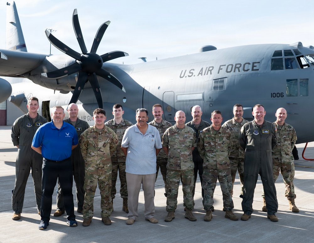 A group of approximately 13 individuals pose for a formal group photograph in front of a U.S. Air Force C-130J Super Hercules aircraft on a flight line or tarmac. The group consists of military personnel in various uniforms including flight suits, OCP (Operational Camouflage Pattern) uniforms, and one individual in civilian attire. The aircraft bears the tail number 1006 and 'U.S. AIR FORCE' markings.