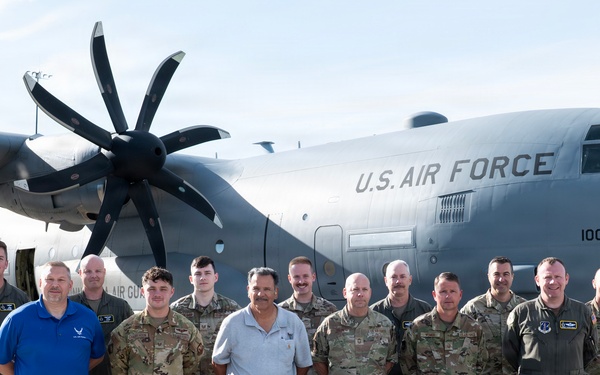 A group of approximately 13 individuals pose for a formal group photograph in front of a U.S. Air Force C-130J Super Hercules aircraft on a flight line or tarmac. The group consists of military personnel in various uniforms including flight suits, OCP (Operational Camouflage Pattern) uniforms, and one individual in civilian attire. The aircraft bears the tail number 1006 and 'U.S. AIR FORCE' markings.