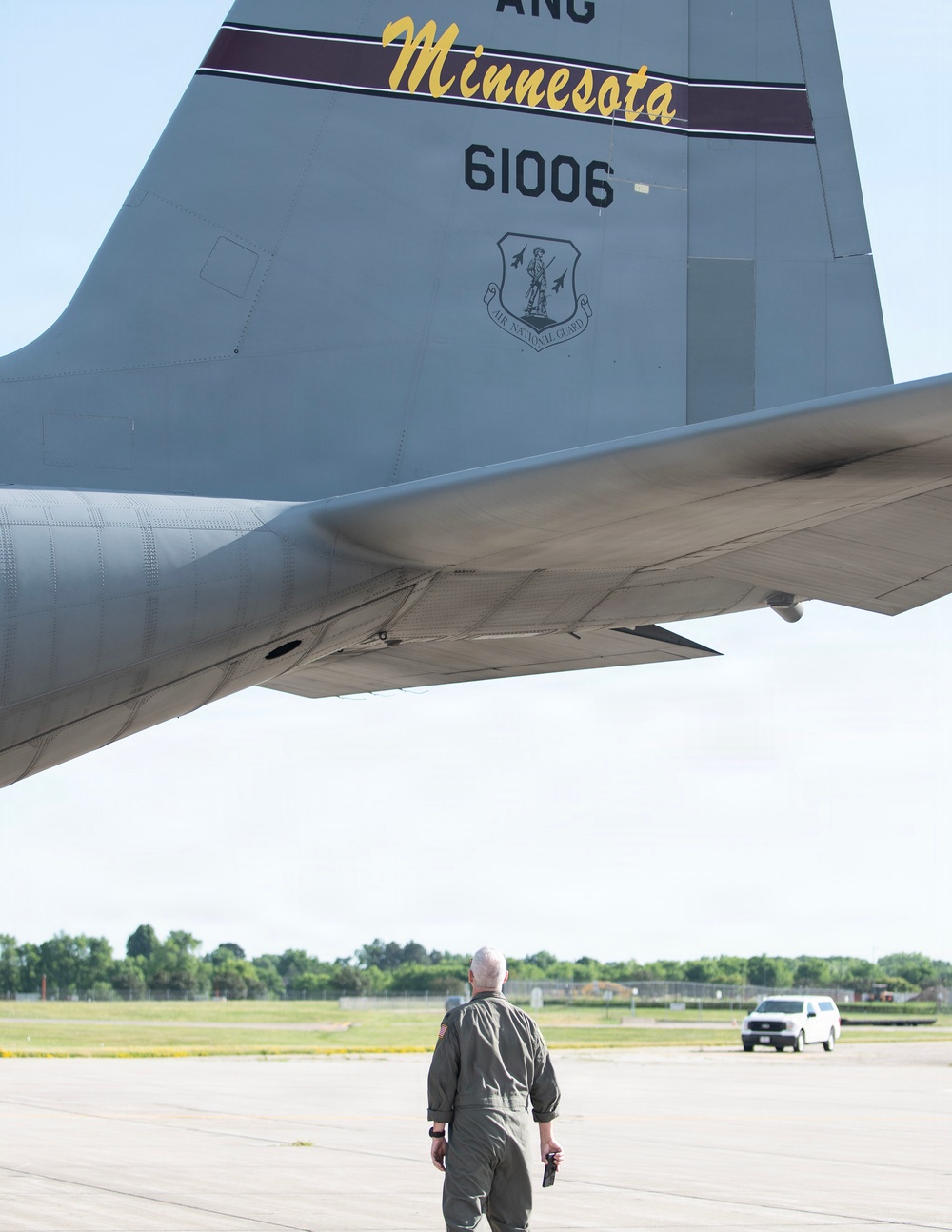 A military aviator in an olive drab flight suit conducts a preflight or post-flight walk-around inspection of a large military transport aircraft on a tarmac. The aircraft's tail section is prominently visible, marked with 'ANG Minnesota,' tail number 61006, and the Air National Guard shield insignia. The setting appears to be a military airfield with trees and a white vehicle visible in the background.