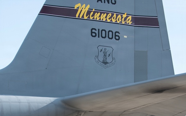 A military aviator in an olive drab flight suit conducts a preflight or post-flight walk-around inspection of a large military transport aircraft on a tarmac. The aircraft's tail section is prominently visible, marked with 'ANG Minnesota,' tail number 61006, and the Air National Guard shield insignia. The setting appears to be a military airfield with trees and a white vehicle visible in the background.