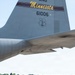 A military aviator in an olive drab flight suit conducts a preflight or post-flight walk-around inspection of a large military transport aircraft on a tarmac. The aircraft's tail section is prominently visible, marked with 'ANG Minnesota,' tail number 61006, and the Air National Guard shield insignia. The setting appears to be a military airfield with trees and a white vehicle visible in the background.