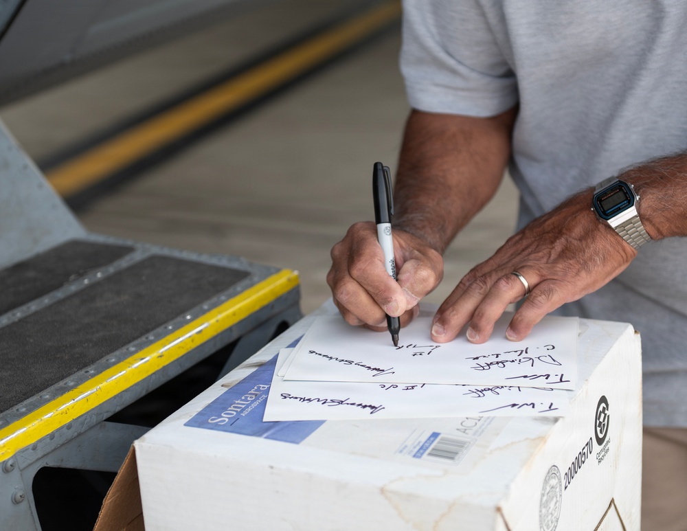 A man is writing on multiple sheets of paper using a black Sharpie marker, using a cardboard box as a writing surface. The papers appear to contain signatures or handwritten names/text. The setting appears to be an industrial or warehouse-type environment.