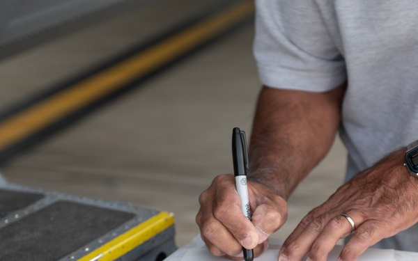 A man is writing on multiple sheets of paper using a black Sharpie marker, using a cardboard box as a writing surface. The papers appear to contain signatures or handwritten names/text. The setting appears to be an industrial or warehouse-type environment.