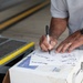 A man is writing on multiple sheets of paper using a black Sharpie marker, using a cardboard box as a writing surface. The papers appear to contain signatures or handwritten names/text. The setting appears to be an industrial or warehouse-type environment.