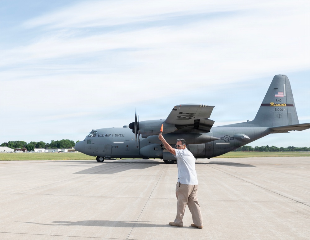 A ground crew member is marshaling a U.S. Air Force C-130 Hercules transport aircraft on a tarmac. The aircraft bears markings identifying it as belonging to the Minnesota Air National Guard (ANG), with tail number 61006. The scene appears to be at a military airfield during daytime under partly cloudy skies.