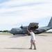 A ground crew member is marshaling a U.S. Air Force C-130 Hercules transport aircraft on a tarmac. The aircraft bears markings identifying it as belonging to the Minnesota Air National Guard (ANG), with tail number 61006. The scene appears to be at a military airfield during daytime under partly cloudy skies.