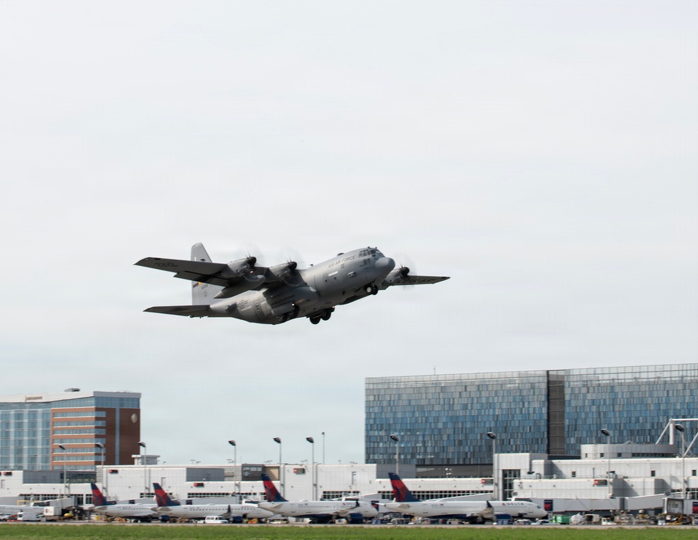 A U.S. Air Force C-130 Hercules military transport aircraft is captured in a low-altitude takeoff or landing at a civilian airport. In the background, multiple Delta Air Lines commercial aircraft are visible at terminal gates, along with airport infrastructure and adjacent buildings. The contrast between the military and civilian aviation assets is a notable feature of this image.