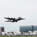 A U.S. Air Force C-130 Hercules military transport aircraft is captured in a low-altitude takeoff or landing at a civilian airport. In the background, multiple Delta Air Lines commercial aircraft are visible at terminal gates, along with airport infrastructure and adjacent buildings. The contrast between the military and civilian aviation assets is a notable feature of this image.