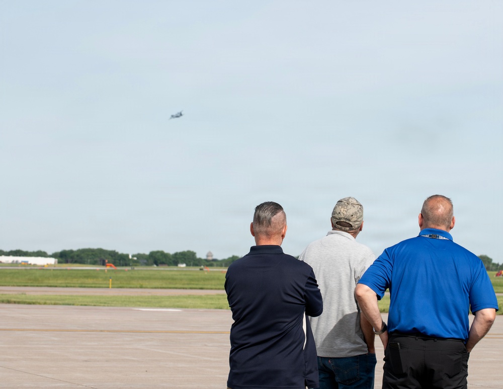 Three men are standing on an airport tarmac or airfield, watching an aircraft flying in the distance. They are viewed from behind, all facing away from the camera toward the sky. The setting appears to be a military or civilian airfield with a flat, open landscape and tree line visible on the horizon.