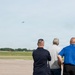 Three men are standing on an airport tarmac or airfield, watching an aircraft flying in the distance. They are viewed from behind, all facing away from the camera toward the sky. The setting appears to be a military or civilian airfield with a flat, open landscape and tree line visible on the horizon.