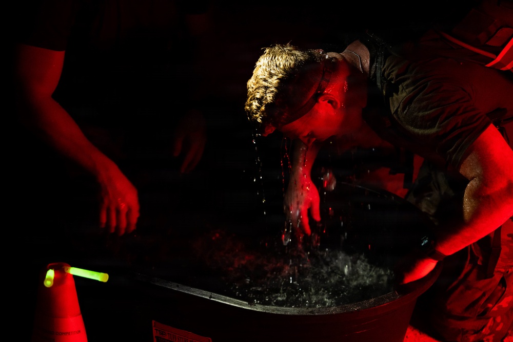 A person is submerging or lifting their head from a large container filled with water, illuminated by intense red lighting in an otherwise dark environment. Water is visibly dripping from their face and hair. A green glow stick and an orange traffic cone are visible in the lower left, suggesting a nighttime event or competition.