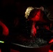 A person is submerging or lifting their head from a large container filled with water, illuminated by intense red lighting in an otherwise dark environment. Water is visibly dripping from their face and hair. A green glow stick and an orange traffic cone are visible in the lower left, suggesting a nighttime event or competition.