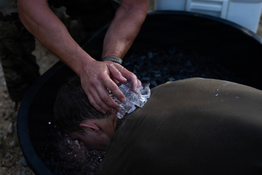 A person wearing an olive/military-colored shirt has their head submerged or lowered into a large black tub filled with ice water. Another individual, wearing a bracelet and camouflage clothing, is applying ice cubes directly to the back of the person's neck and head. This appears to be a heat casualty treatment or cold water immersion procedure, consistent with military medical protocols for heat stroke management.
