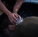A person wearing an olive/military-colored shirt has their head submerged or lowered into a large black tub filled with ice water. Another individual, wearing a bracelet and camouflage clothing, is applying ice cubes directly to the back of the person's neck and head. This appears to be a heat casualty treatment or cold water immersion procedure, consistent with military medical protocols for heat stroke management.