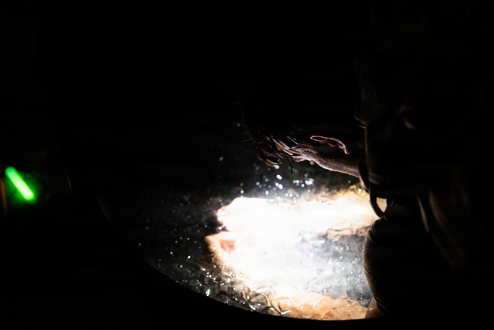 A dark, low-light photograph showing a hand reaching over what appears to be a brightly illuminated surface, possibly a cooking vessel or pan with bubbling liquid. The scene is dramatically backlit with a single intense light source creating strong contrast against the near-total darkness. A small green light is visible on the left side of the frame.