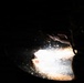 A dark, low-light photograph showing a hand reaching over what appears to be a brightly illuminated surface, possibly a cooking vessel or pan with bubbling liquid. The scene is dramatically backlit with a single intense light source creating strong contrast against the near-total darkness. A small green light is visible on the left side of the frame.