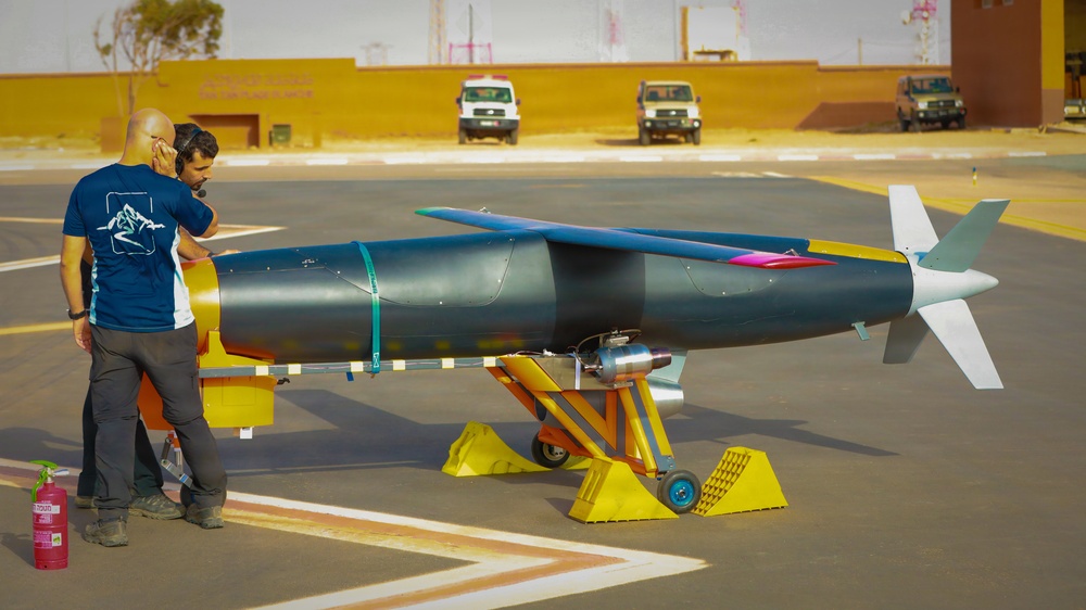 Two technicians are inspecting a large rocket or missile-shaped vehicle mounted on a yellow wheeled support cradle on a tarmac surface. The vehicle appears to be a test article or experimental aerospace craft with colorful markings, fins at the rear, and a jet engine visible underneath. The setting appears to be a desert test facility, with military or utility vehicles visible in the background.