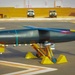 Two technicians are inspecting a large rocket or missile-shaped vehicle mounted on a yellow wheeled support cradle on a tarmac surface. The vehicle appears to be a test article or experimental aerospace craft with colorful markings, fins at the rear, and a jet engine visible underneath. The setting appears to be a desert test facility, with military or utility vehicles visible in the background.