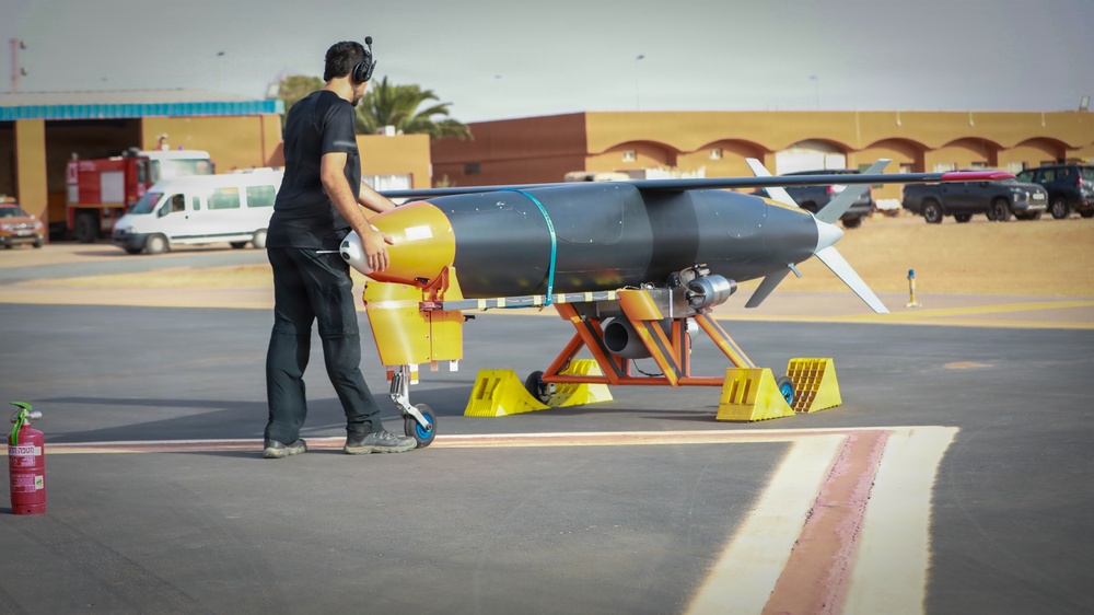 A technician in black clothing and hearing protection is working on a large unmanned aerial vehicle (UAV) or target drone mounted on a yellow ground support stand on an airfield tarmac. The aircraft has a dark gray cylindrical fuselage with swept wings and a rear-mounted engine. The setting appears to be a military or test airfield in an arid environment.