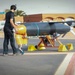 A technician in black clothing and hearing protection is working on a large unmanned aerial vehicle (UAV) or target drone mounted on a yellow ground support stand on an airfield tarmac. The aircraft has a dark gray cylindrical fuselage with swept wings and a rear-mounted engine. The setting appears to be a military or test airfield in an arid environment.