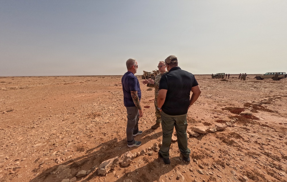 Three men are standing in a arid desert environment engaged in conversation. One individual appears to be wearing military camouflage attire, while the other two are in civilian clothing. In the background, military vehicles, personnel, and equipment are visible on the flat desert terrain.