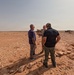 Three men are standing in a arid desert environment engaged in conversation. One individual appears to be wearing military camouflage attire, while the other two are in civilian clothing. In the background, military vehicles, personnel, and equipment are visible on the flat desert terrain.