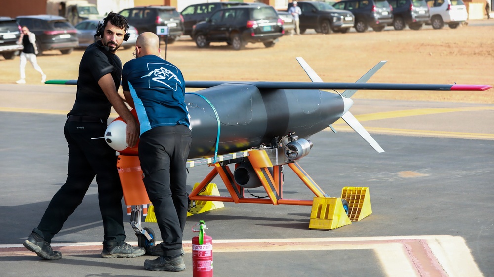 Two technicians are working on a jet-powered unmanned aerial vehicle (UAV) or target drone mounted on an orange launch stand with yellow wheel chocks. The aircraft has a torpedo-shaped fuselage, straight wings with colored wingtips, and a rear-mounted jet engine. The setting appears to be an outdoor airfield or demonstration area, likely at a defense or aerospace exhibition.