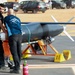 Two technicians are working on a jet-powered unmanned aerial vehicle (UAV) or target drone mounted on an orange launch stand with yellow wheel chocks. The aircraft has a torpedo-shaped fuselage, straight wings with colored wingtips, and a rear-mounted jet engine. The setting appears to be an outdoor airfield or demonstration area, likely at a defense or aerospace exhibition.