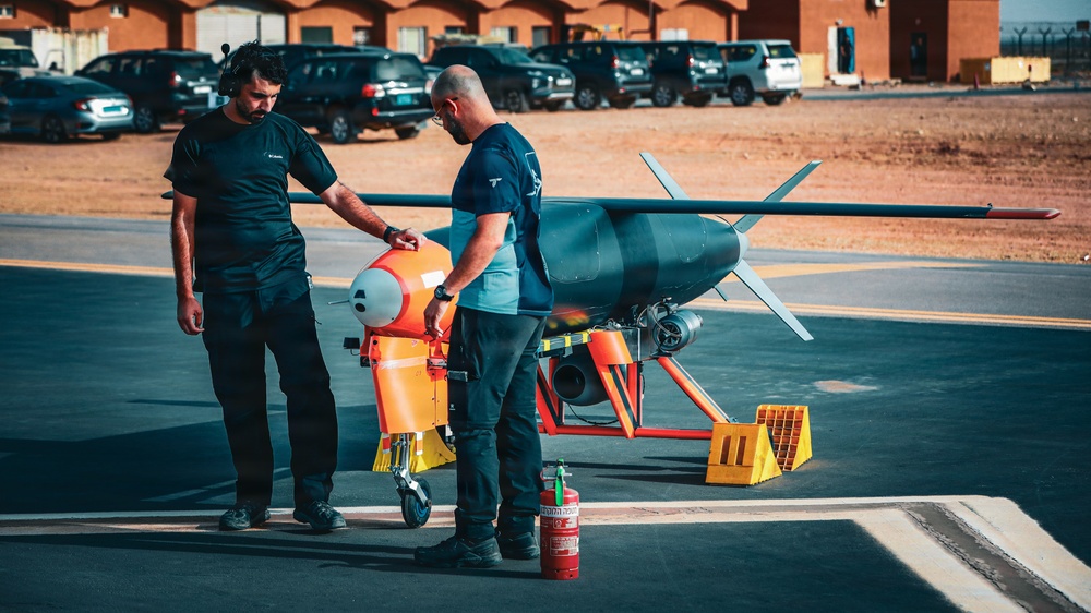 Two technicians are conducting pre-flight or maintenance checks on an unmanned aerial vehicle (UAV) or target drone on a tarmac. The aircraft features a distinctive orange and gray color scheme with a pusher propeller configuration. A fire extinguisher is positioned nearby as a safety precaution, and several vehicles and a building are visible in the background.