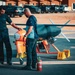 Two technicians are conducting pre-flight or maintenance checks on an unmanned aerial vehicle (UAV) or target drone on a tarmac. The aircraft features a distinctive orange and gray color scheme with a pusher propeller configuration. A fire extinguisher is positioned nearby as a safety precaution, and several vehicles and a building are visible in the background.