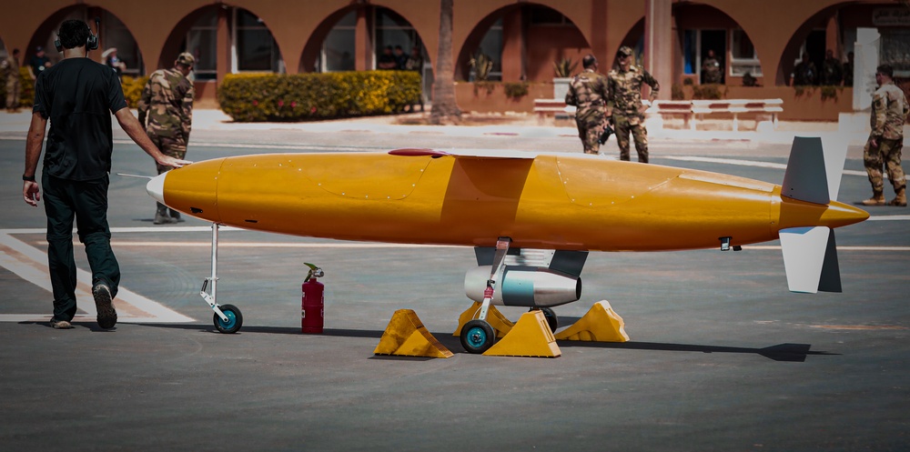 The image shows a yellow unmanned aerial vehicle (UAV) or target drone on a tarmac or courtyard surface, secured with wheel chocks and supported by landing gear. Military personnel in camouflage uniforms are visible in the background near an arched building, suggesting a military demonstration or exercise. A civilian technician in black clothing is walking alongside the aircraft.