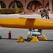 The image shows a yellow unmanned aerial vehicle (UAV) or target drone on a tarmac or courtyard surface, secured with wheel chocks and supported by landing gear. Military personnel in camouflage uniforms are visible in the background near an arched building, suggesting a military demonstration or exercise. A civilian technician in black clothing is walking alongside the aircraft.