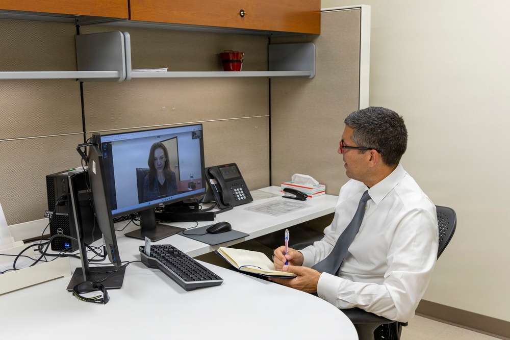 A professional man in a white dress shirt and gray tie sits at an office cubicle desk conducting a video call with a woman displayed on a Dell monitor. He is taking notes in an open notebook while holding a pen, suggesting a formal meeting or interview setting. The workspace is clean and organized with standard office equipment visible.
