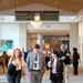Three conference attendees wearing lanyards and name badges walk together through a large hotel convention center corridor, engaged in conversation. The venue appears to be a upscale hotel with a vaulted wood-beam ceiling, chandelier, and directional signage indicating ballroom-level meeting rooms. Additional conference attendees are visible in the background.
