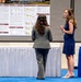 Two women are engaged in conversation at what appears to be an academic or military research poster session. One woman faces away from the camera wearing a gray suit, while the other faces forward wearing a navy blue dress and glasses. The research posters visible in the background relate to military training and nutrition topics, with a DHA (Defense Health Agency) logo visible.