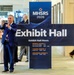 A man in a blue suit is taking a selfie next to a large cross-shaped display sign for MHSRS 2026 Exhibit Hall at what appears to be a conference or convention center. The sign displays Exhibit Hall hours for Monday August 3, Tuesday August 4, and Wednesday August 5. Several other conference attendees are visible in the background.