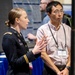 A U.S. Army officer in dress uniform, identified by name tag as Megan McDermott, is presenting or explaining something using hand gestures at what appears to be an academic or professional conference. She is speaking to a man wearing a conference badge identifying him as Lizhi Wang from George Mason University. The setting appears to be a poster session or exhibition hall with research displays visible in the background.