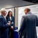 A professional academic or medical conference poster session is taking place in a convention center. Three individuals in business attire are engaged in conversation in front of a research poster about 'Adrenergic-Induced Cardiac Action Potential Shortening' from USU (Uniformed Services University). In the background, additional attendees are visible, including one person wearing a kilt and colorful mohawk hairstyle.