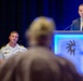 A formal conference or event is taking place in a large venue with dark blue curtain backdrop. A man in a navy blue suit is speaking at a illuminated blue podium bearing a sun/gear logo, while a uniformed military officer (appearing to be U.S. Navy, Captain rank based on visible insignia) stands nearby. In the foreground, an out-of-focus audience member is visible with raised hands, suggesting a Q&amp;A session.