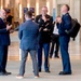 Four men are engaged in a lively conversation in what appears to be a large conference center or government building lobby. Two of the men are wearing military uniforms — one in U.S. Army dress blues and one in U.S. Air Force dress blues — while the other two are in civilian business attire. The group appears to be sharing a humorous moment, with visible laughter and animated gestures.